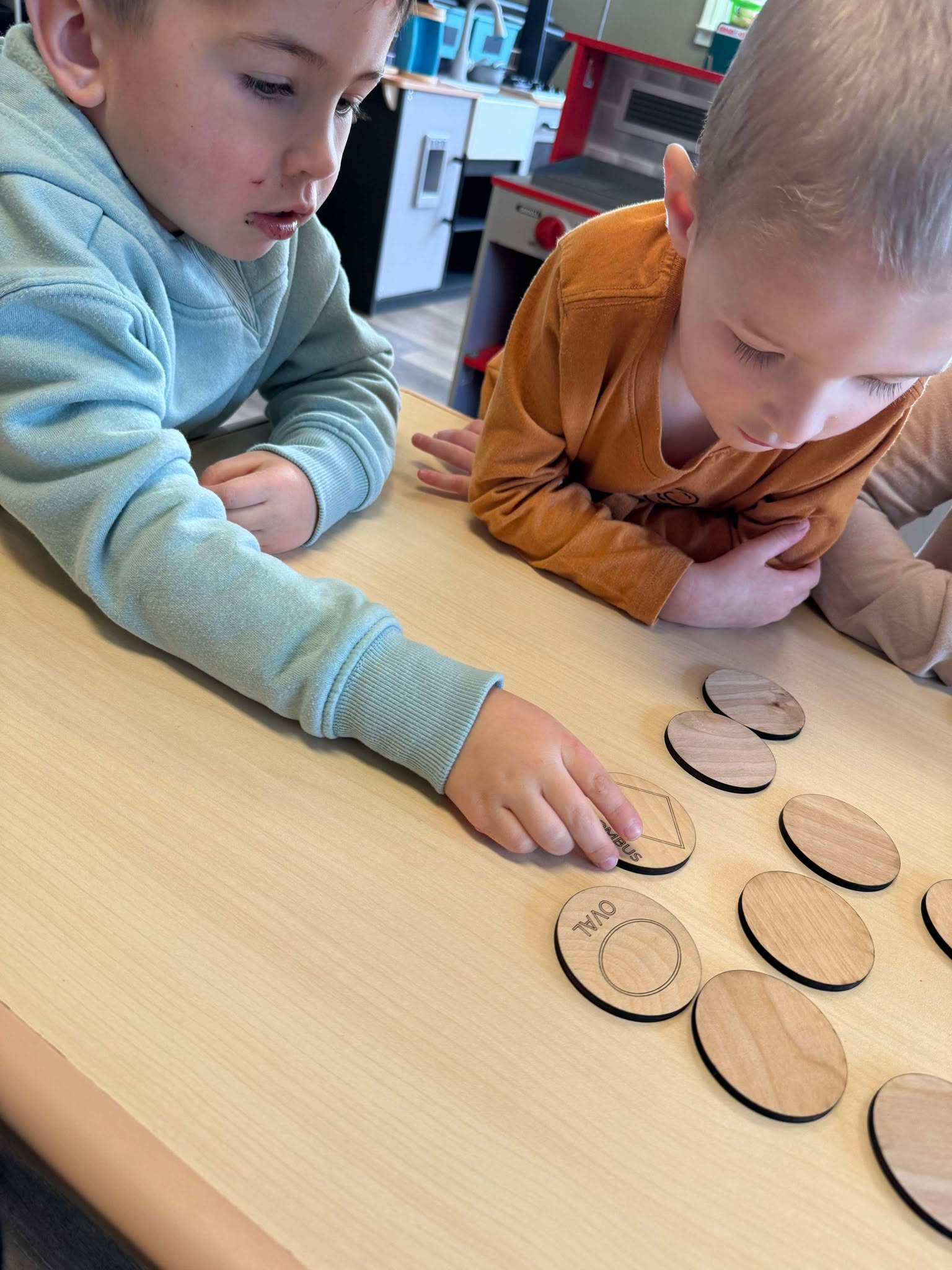 Two boys collaborating on the Basic Shapes Matching Game, fostering social skills and problem solving in a daycare setting while matching labeled wooden tiles like ‘Oval’ and ‘Rhombus'.